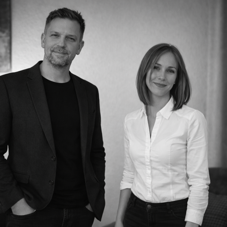 Zwei Personen stehen nebeneinander, der Mann trägt einen dunklen Blazer, die Frau ein weißes Hemd.